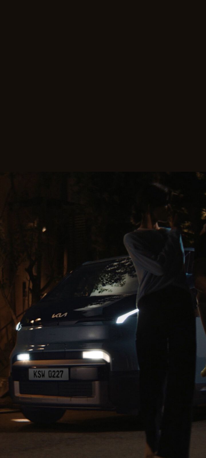 Nächtliche Szene: Der Kia PV5 Passenger mit eingeschaltetem Licht, während eine Familie davor steht.
